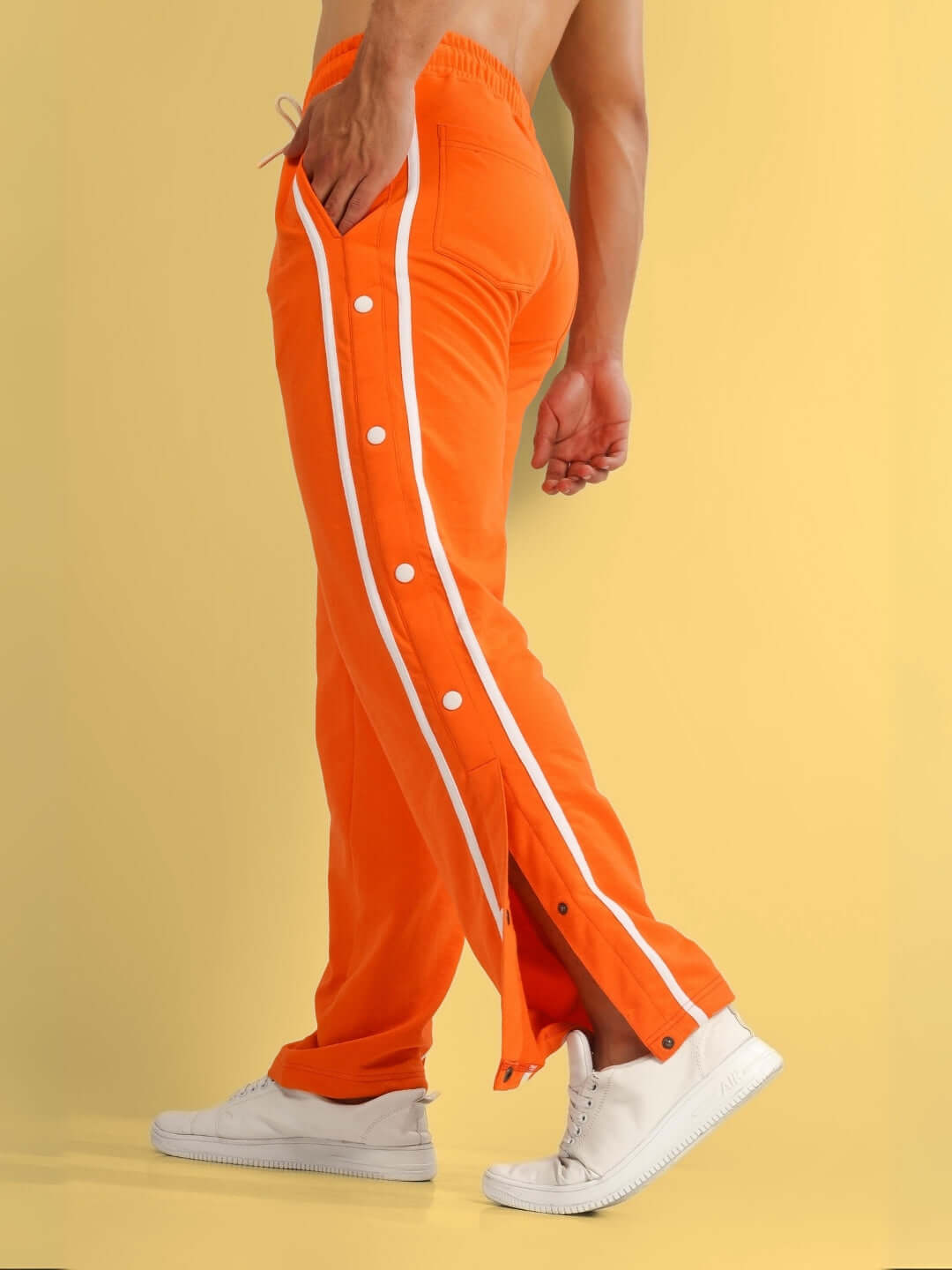 Model wearing orange contrast panel jogger pants with white sneakers showcasing side buttons against a yellow background.