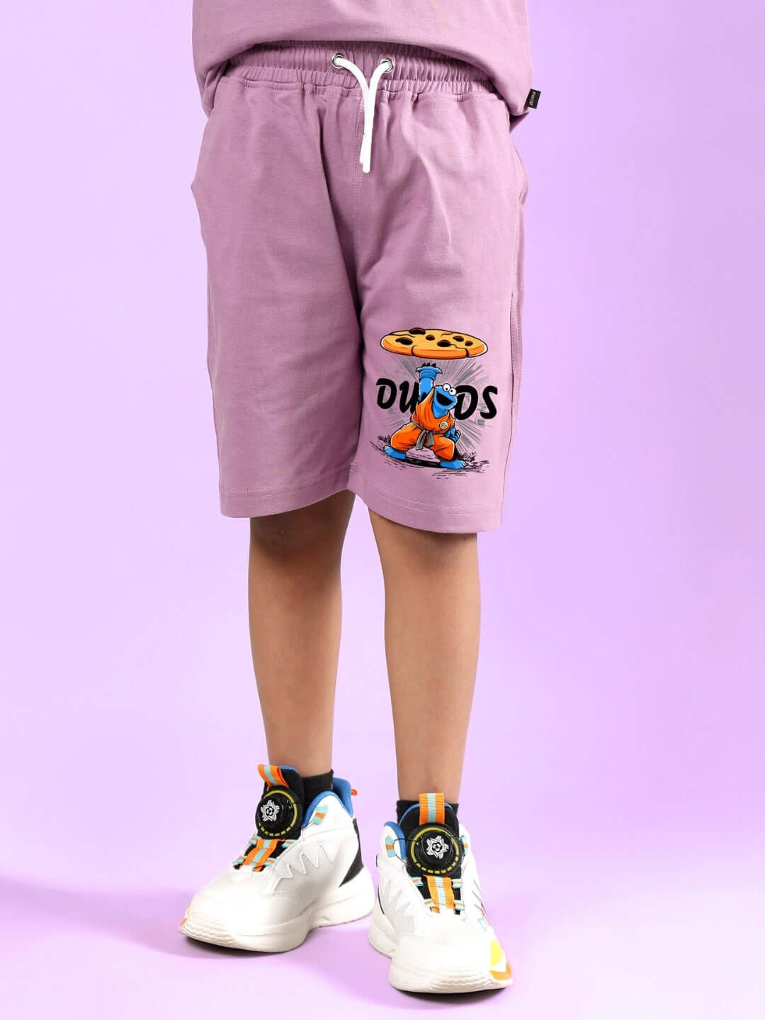 Child wearing COOKIE MONSTER PRINTED CO-ORD FOR BOYS & GIRLS PLUM PURPLE shorts with DUDS logo on a purple background.