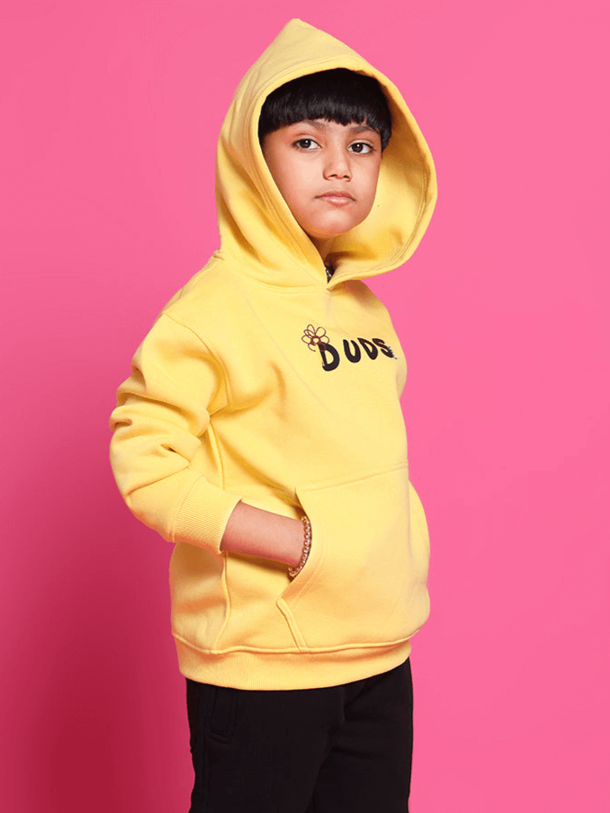 Child wearing yellow oversized COOL DUDE hoodie with hands in pockets, standing against pink background.