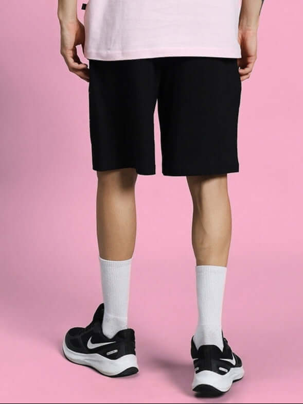 Person wearing COOL PALS REGULAR FIT SHORTS BLACK on pink background with white socks and sneakers.