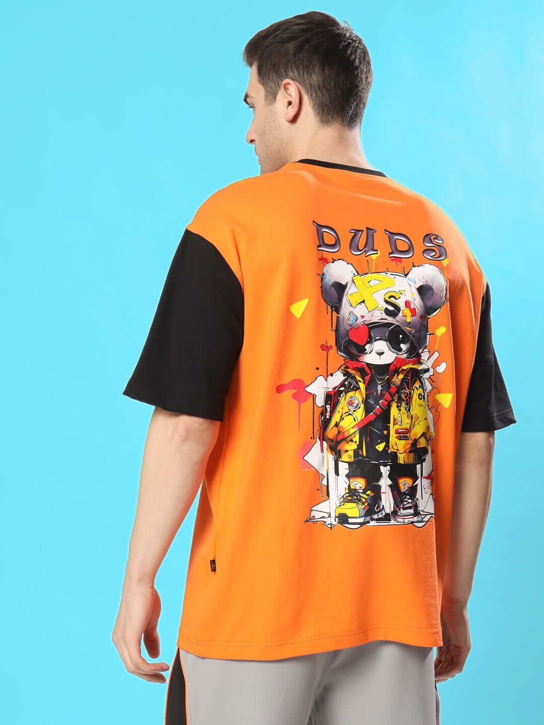 Back view of a man wearing a COOL TEDDY oversized orange-black t-shirt, featuring a stylish teddy graphic on a vibrant blue background.