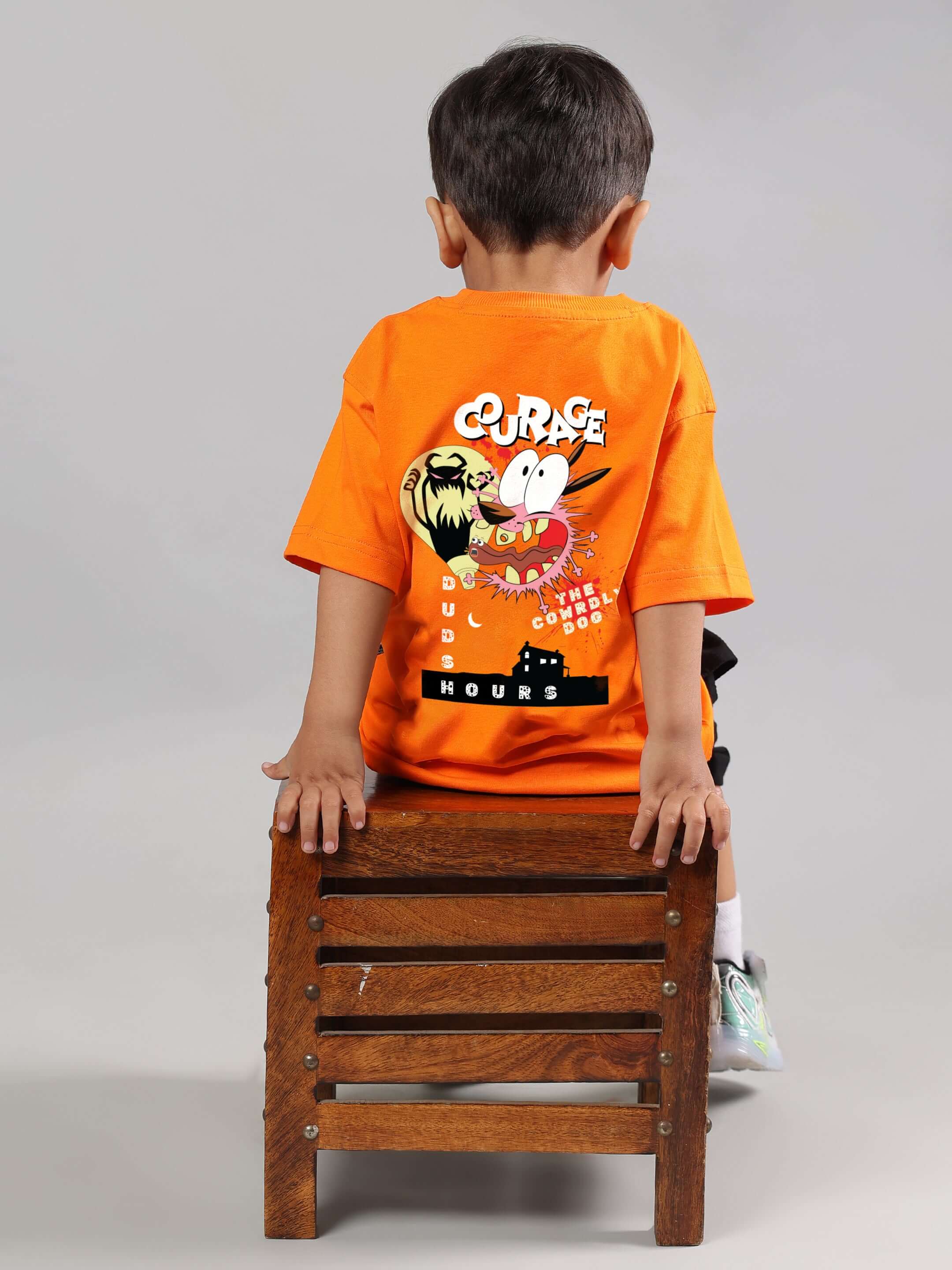 Child wearing an orange Courage Toon T-Shirt for boys and girls, sitting on a wooden chair, showcasing the vibrant cartoon design on the back.