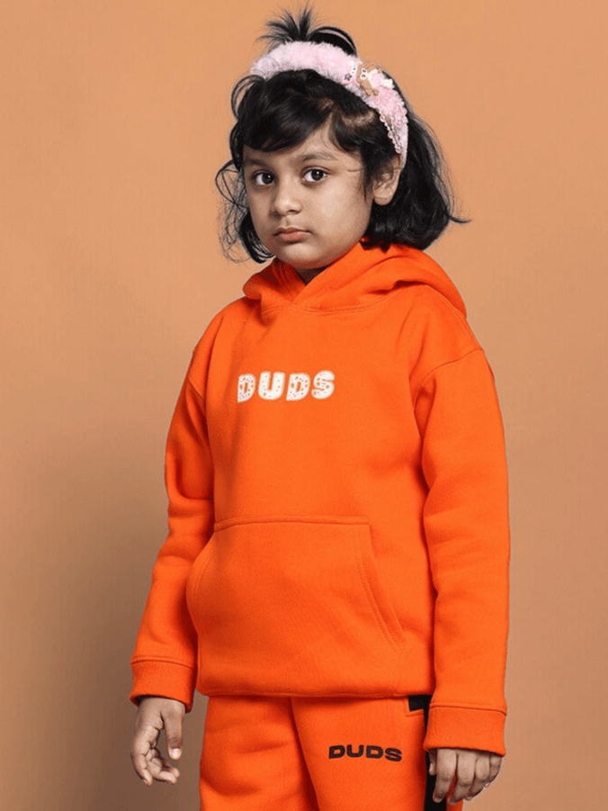 Young child wearing COURAGE OVERSIZED HOODIE BOYS & GIRLS ORANGE with text 'DUDS' on a warm background. Cozy and vibrant clothing for kids.
