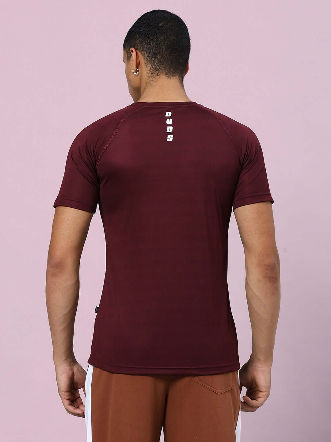 Back view of a man wearing a maroon CRISTINIO regular fit t-shirt made from sports polyester fabric, standing against a pink background.