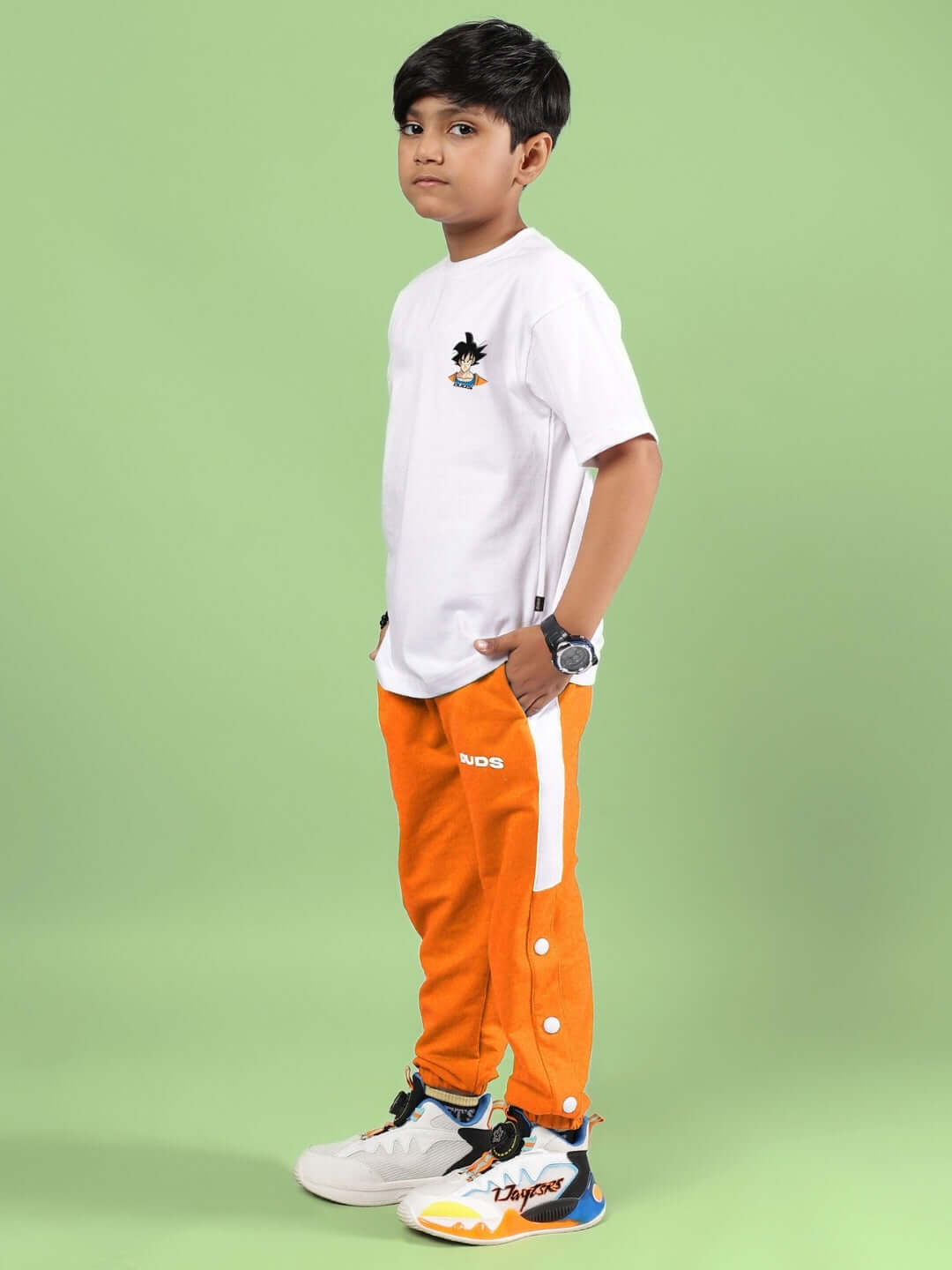 Boy wearing CROSSOVER CO-ORD FOR BOYS GIRLS WHITE-ORANGE featuring white tee and orange joggers, standing on green background.