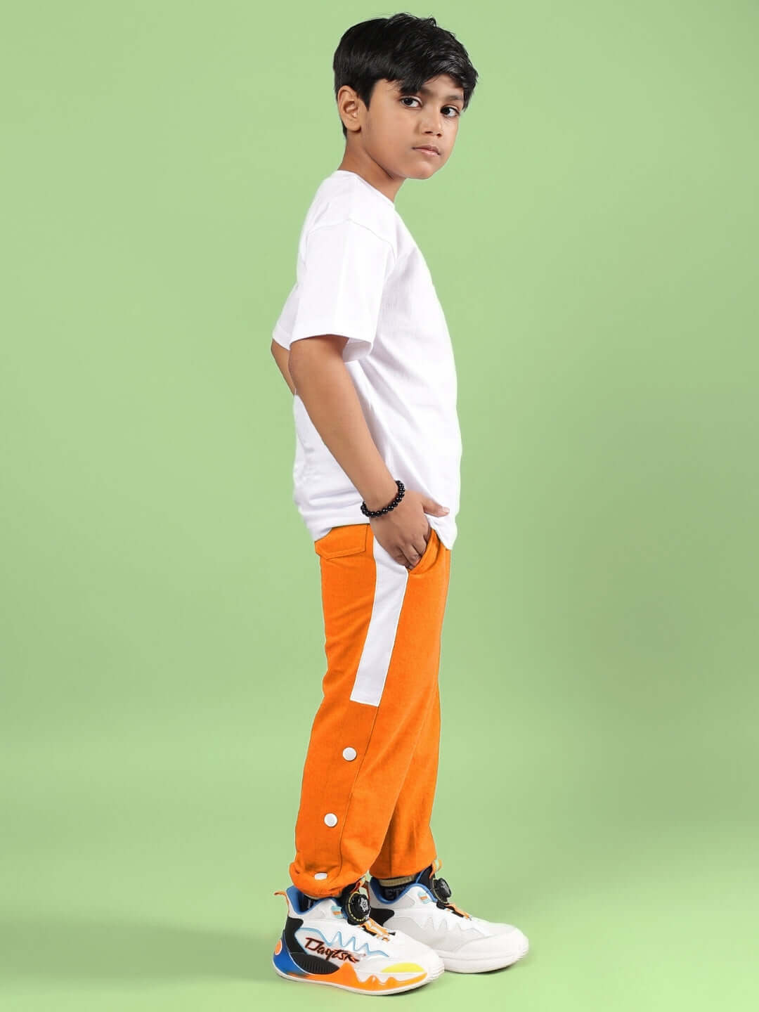 Child wearing CROSSOVER CO-ORD FOR BOYS GIRLS WHITE-ORANGE, featuring a white T-shirt and orange joggers, standing against a green background.