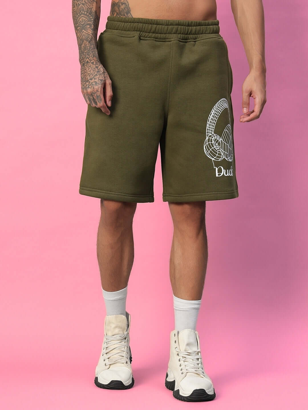 Man wearing OPERA FLEECE SHORTS OLIVE GREEN with white sneakers on pink background