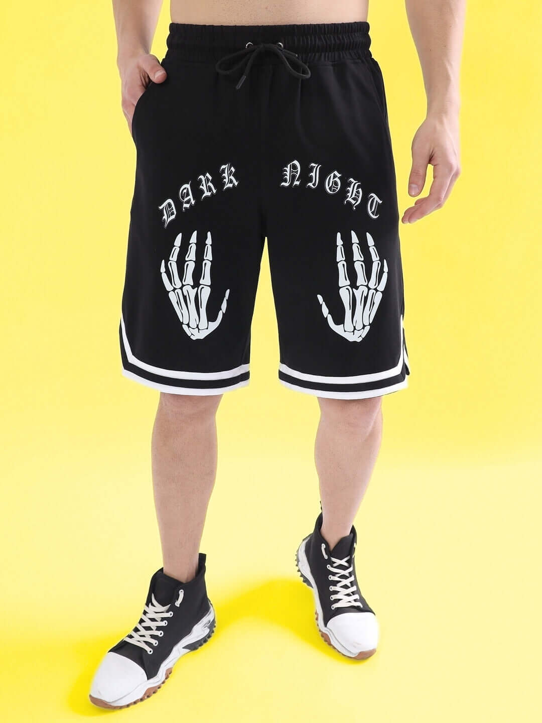 Model wearing Dark Night regular fit black shorts with skeleton hand design, showcasing urban fashion style on a yellow background.