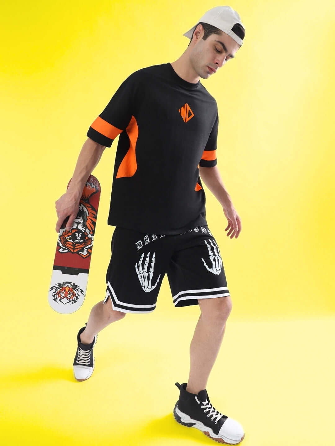 Man wearing DARK NIGHT Regular Fit Shorts in black, paired with a black and orange shirt, holding a skateboard, against a yellow background.