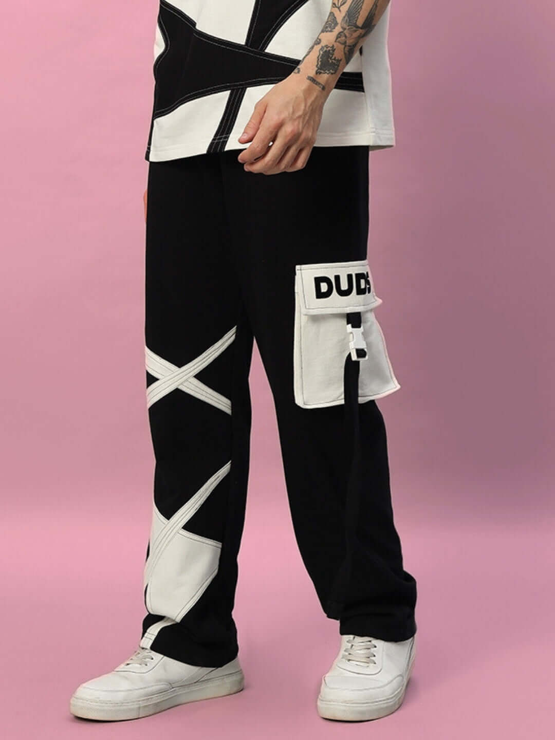 Person wearing black DAVID JOGGER with white cross patterns and a patch pocket, styled with casual sneakers against a pink background.