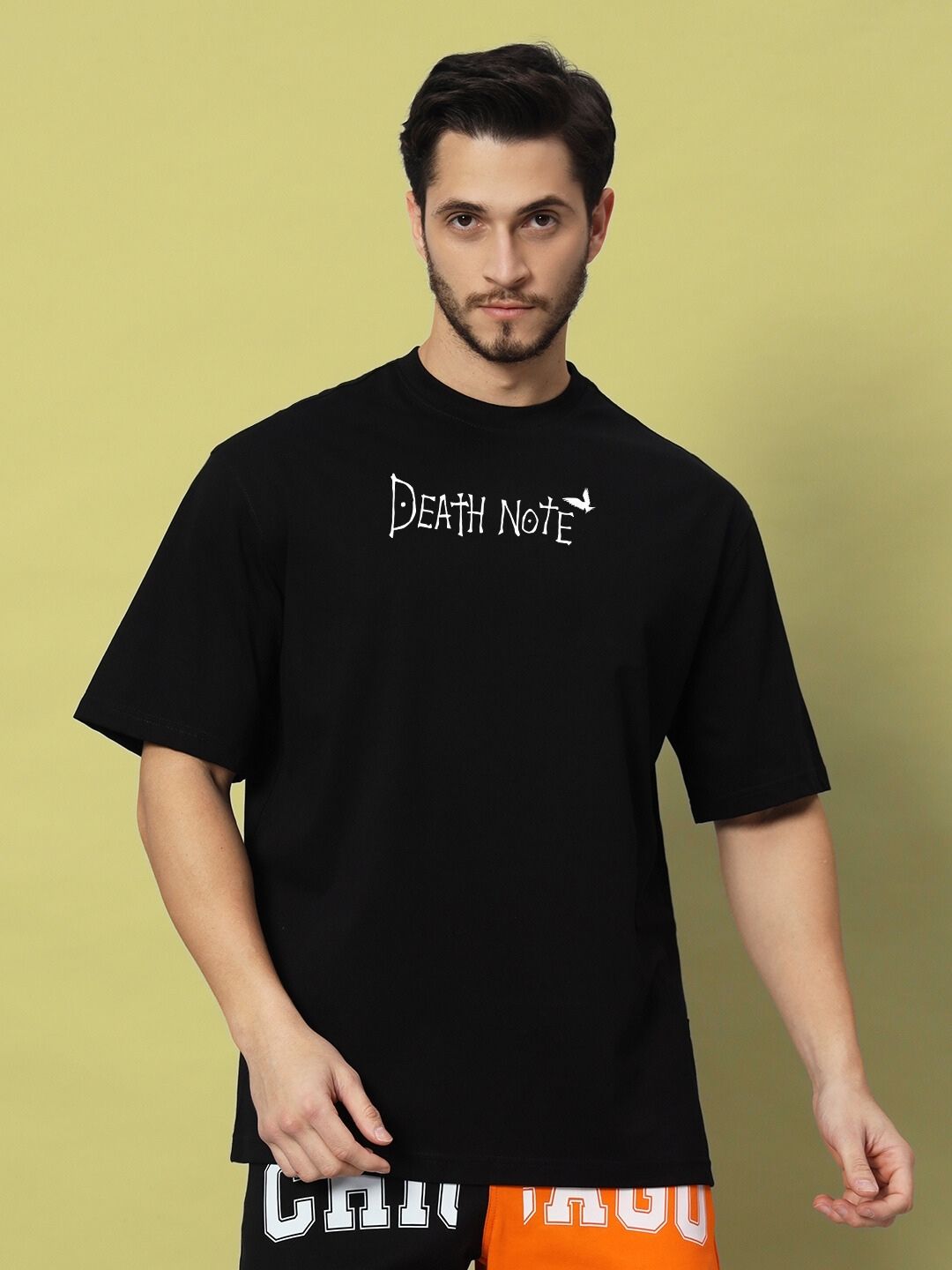 Man wearing FOLLOW LIFE OVERSIZED T-SHIRT BLACK with Death Note design on front, standing against a yellow background.