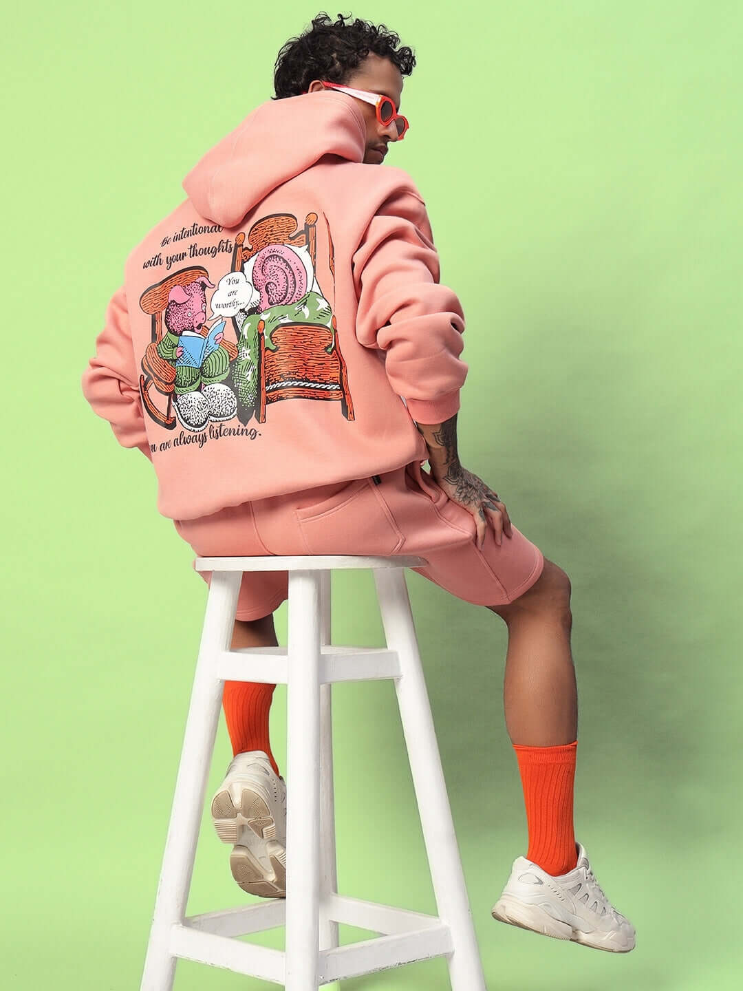 Man wearing DELIBERATE FLEECE CO-ORD PEACH loungewear sitting on stool with artistic hoodie design, green background.