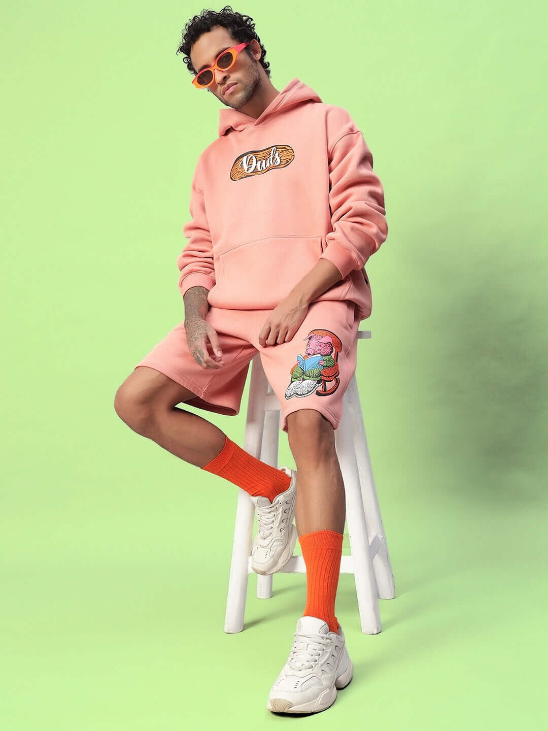 Man wearing DELIBERATE FLEECE CO-ORD PEACH loungewear set, featuring hoodie and shorts on a green background.