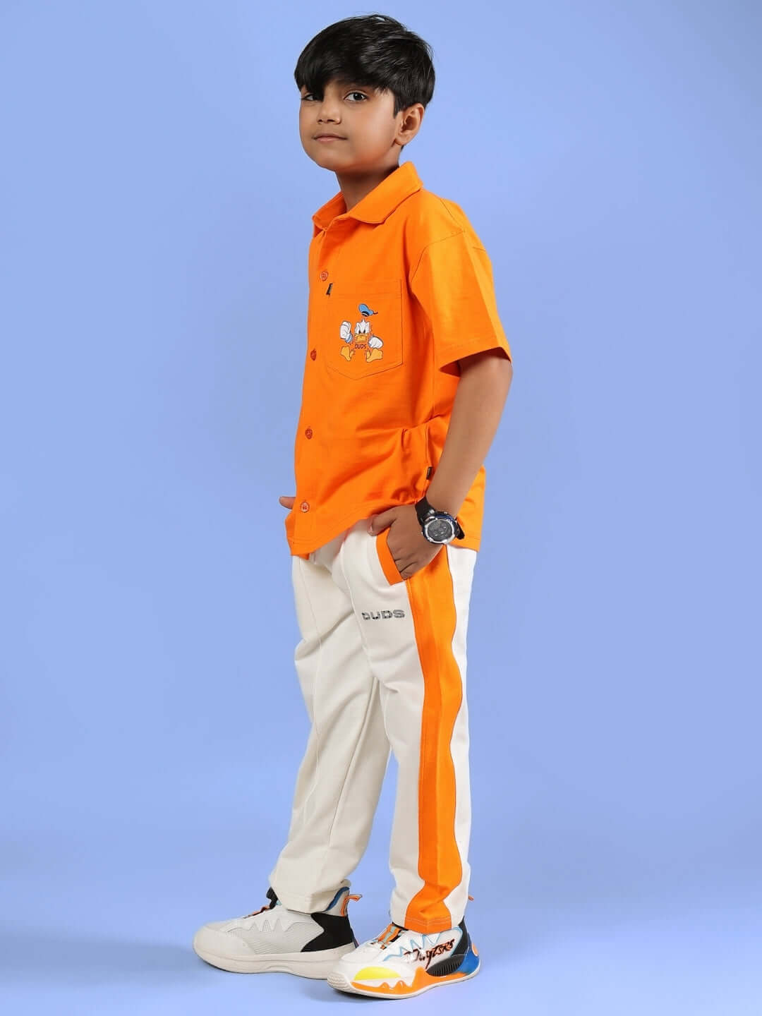 Boy wearing DELLA CO-ORD FOR BOYS & GIRLS ORANGE with sporty style, white pants, and sneakers, set against a blue background.