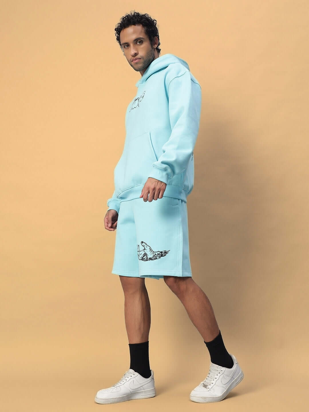 Man wearing WEARDUDS DEMON FLEECE CO-ORD SKY BLUE, featuring a hoodie and shorts, standing against a beige background, showcasing casual style.