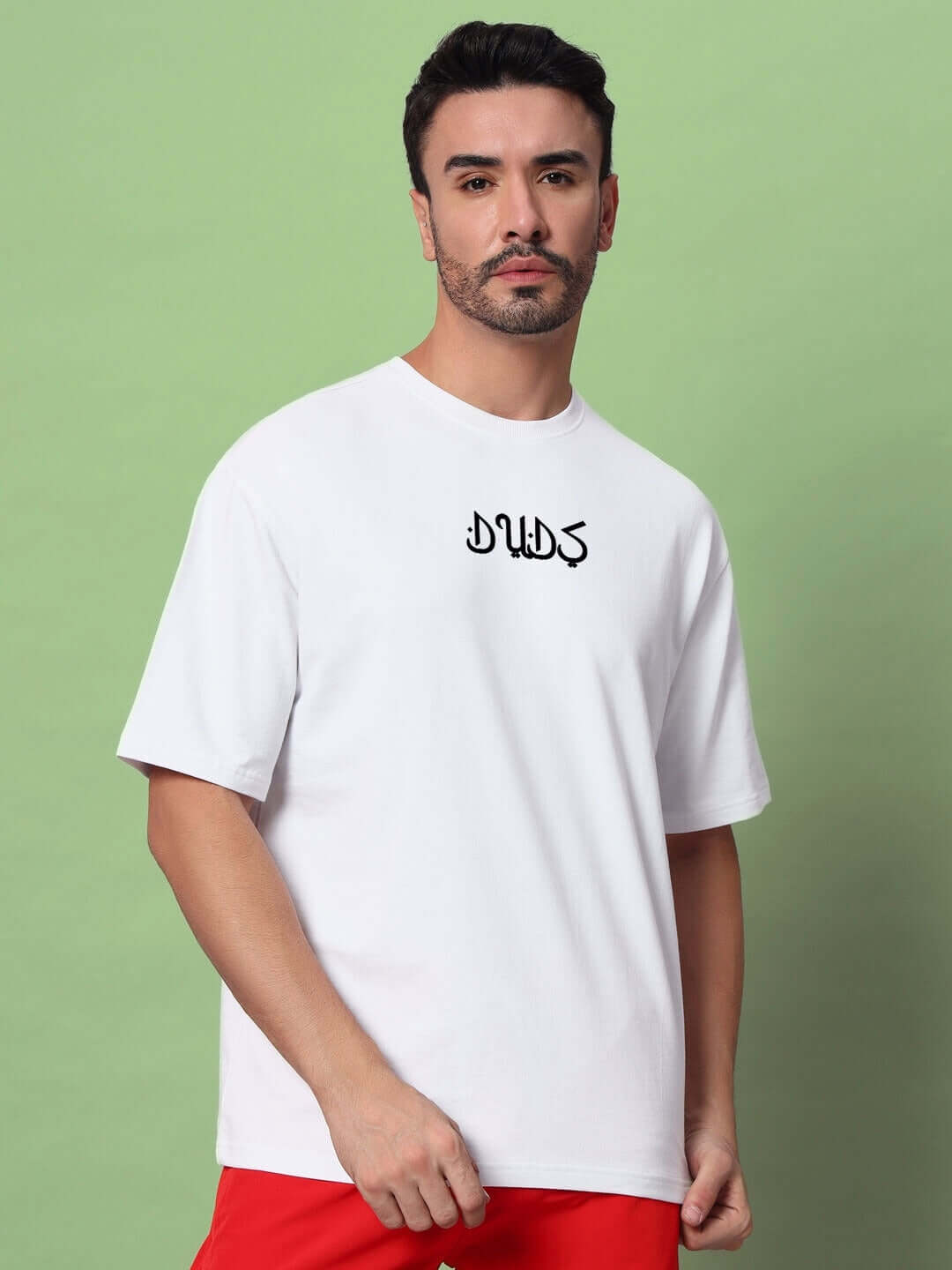 Man wearing DIMENSION oversized white t-shirt made of premium heavy gauge fabric, featuring DUDS logo, on a green background.