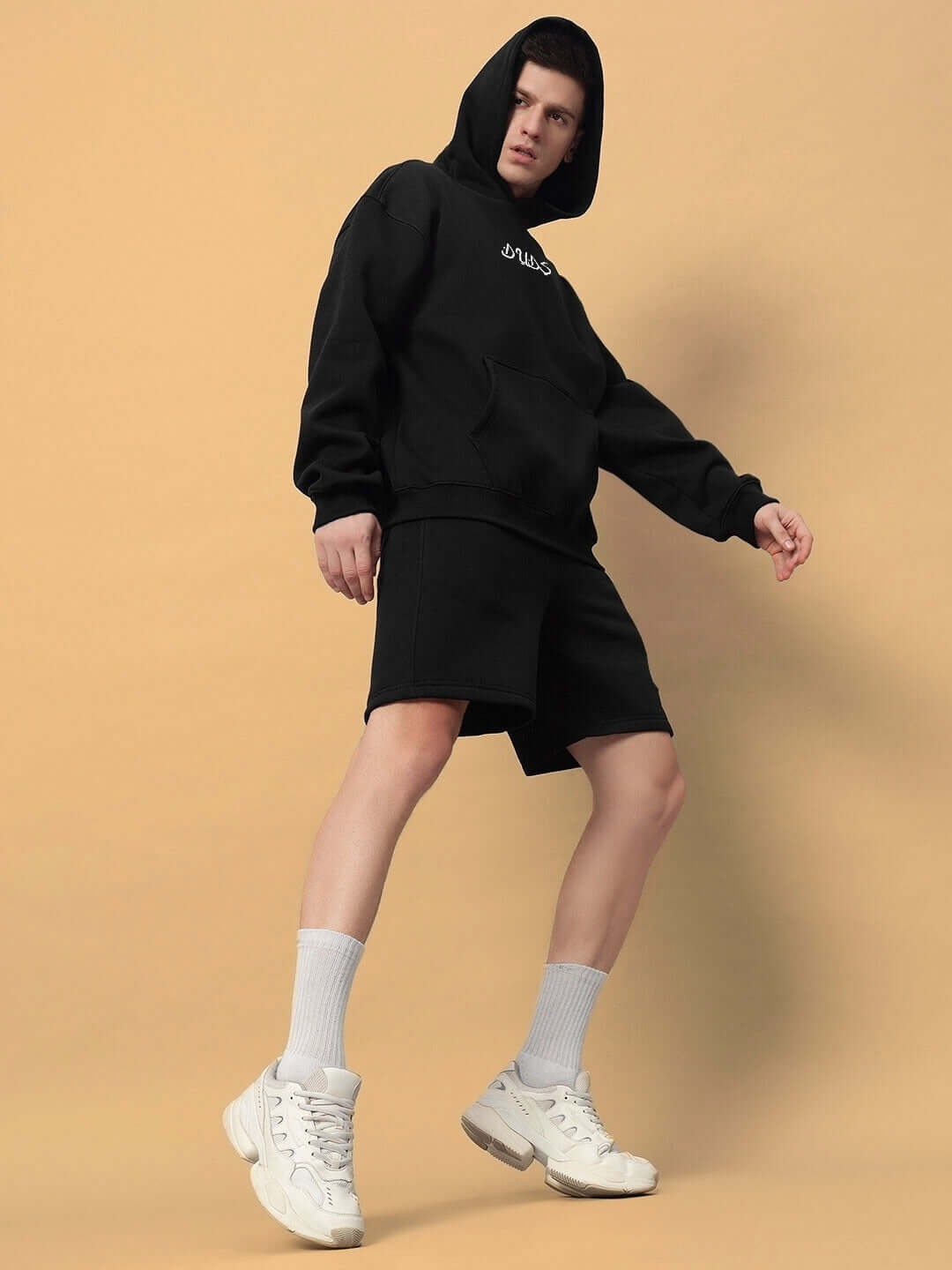 Youth wearing DIMENSION COTTON FLEECE CO-ORD BLACK set, featuring a hoodie and shorts, standing stylishly in front of a beige background.