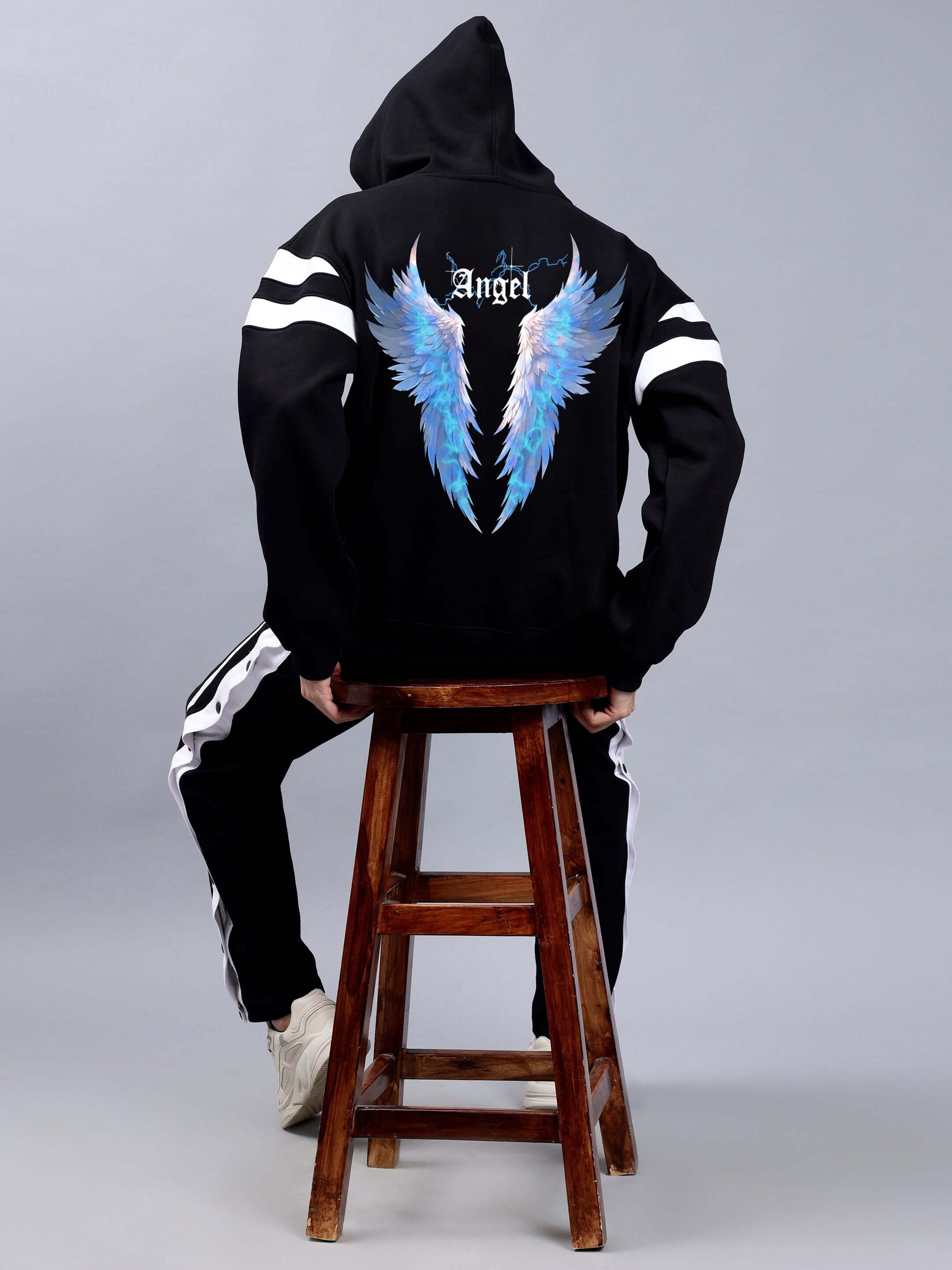 Back view of black colorblock oversized hoodie with angel wing design, worn by model seated on stool.