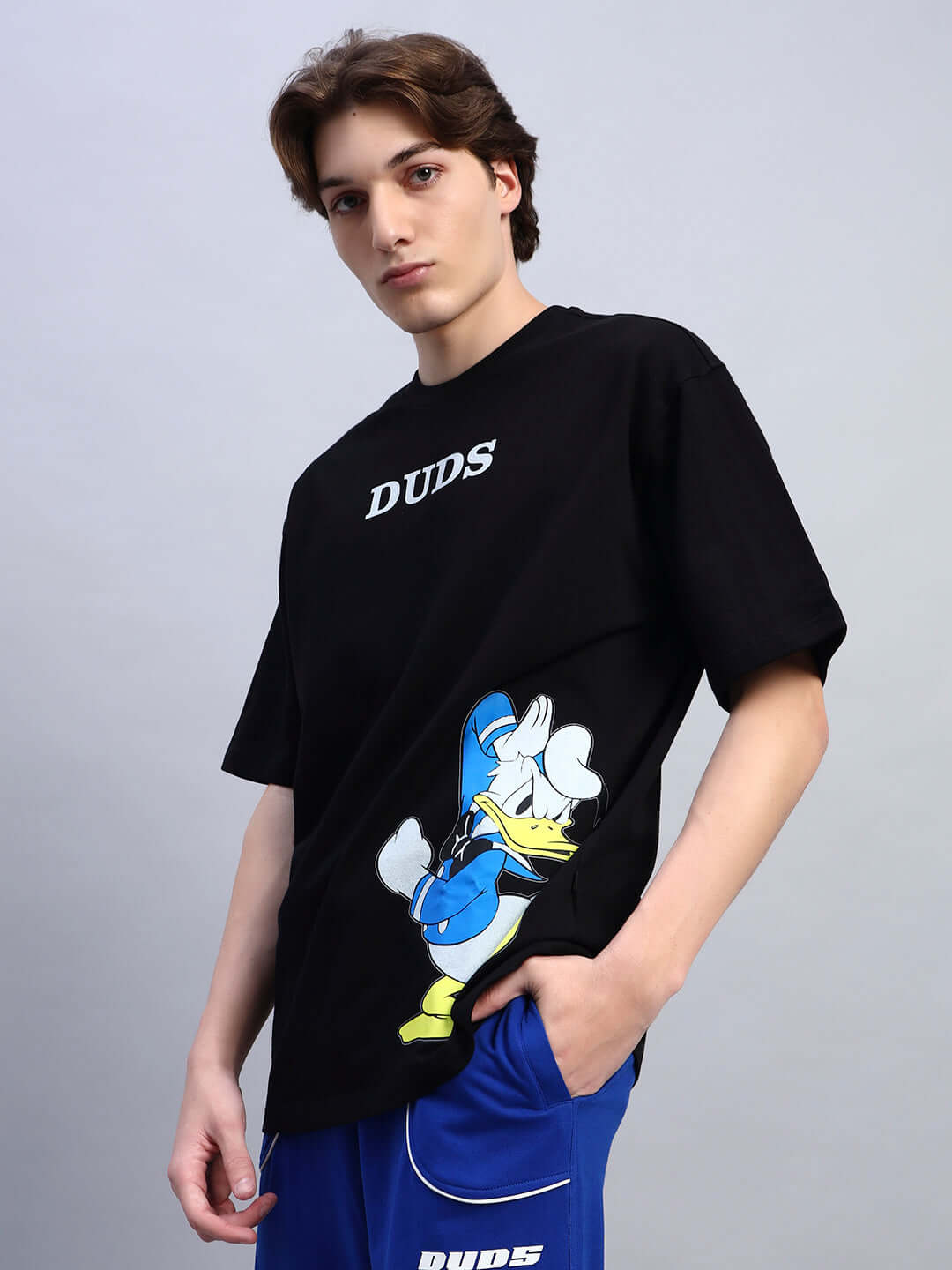 Black oversized Donald Duck printed t-shirt, made from premium fabric, worn by a model in blue pants. Vibrant cartoon graphic design.