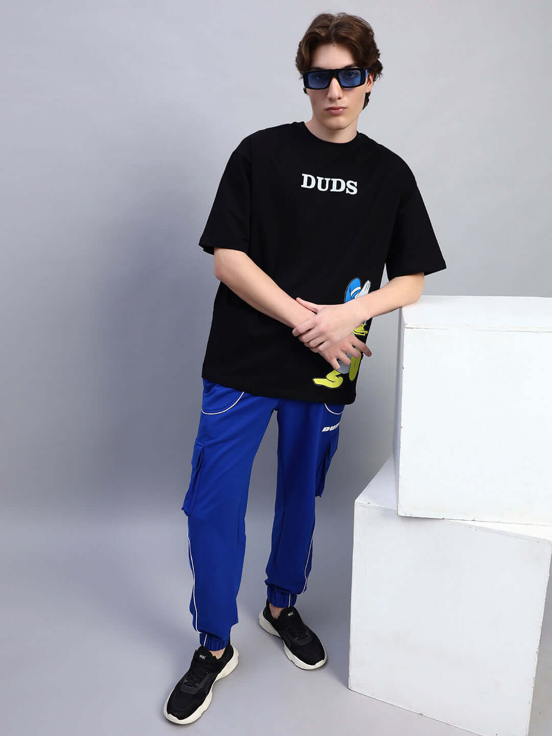 Model wearing DONALD DUCK PRINTED OVERSIZED T-SHIRT BLACK with blue pants and sunglasses, showcasing relaxed streetwear fashion.