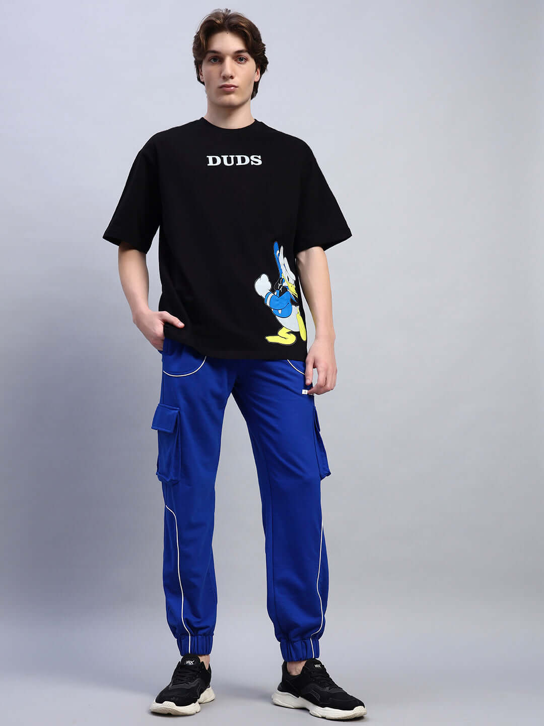 Model wearing DONALD DUCK PRINTED OVERSIZED T-SHIRT BLACK with bold graphic, paired with blue cargo pants, showcasing streetwear style.