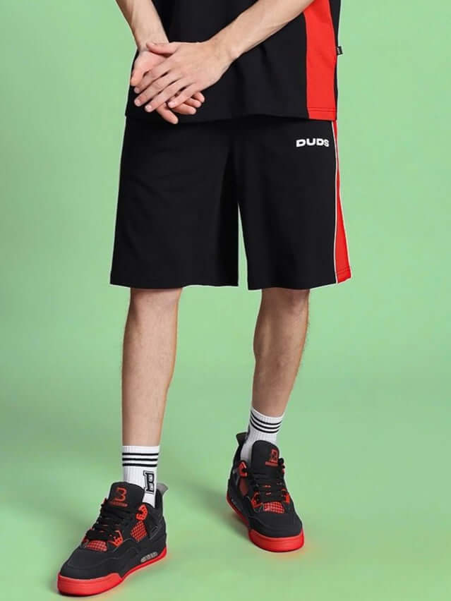 Person wearing DRIFT REGULAR FIT COTTON SHORTS BLACK with red and black sneakers on green background.