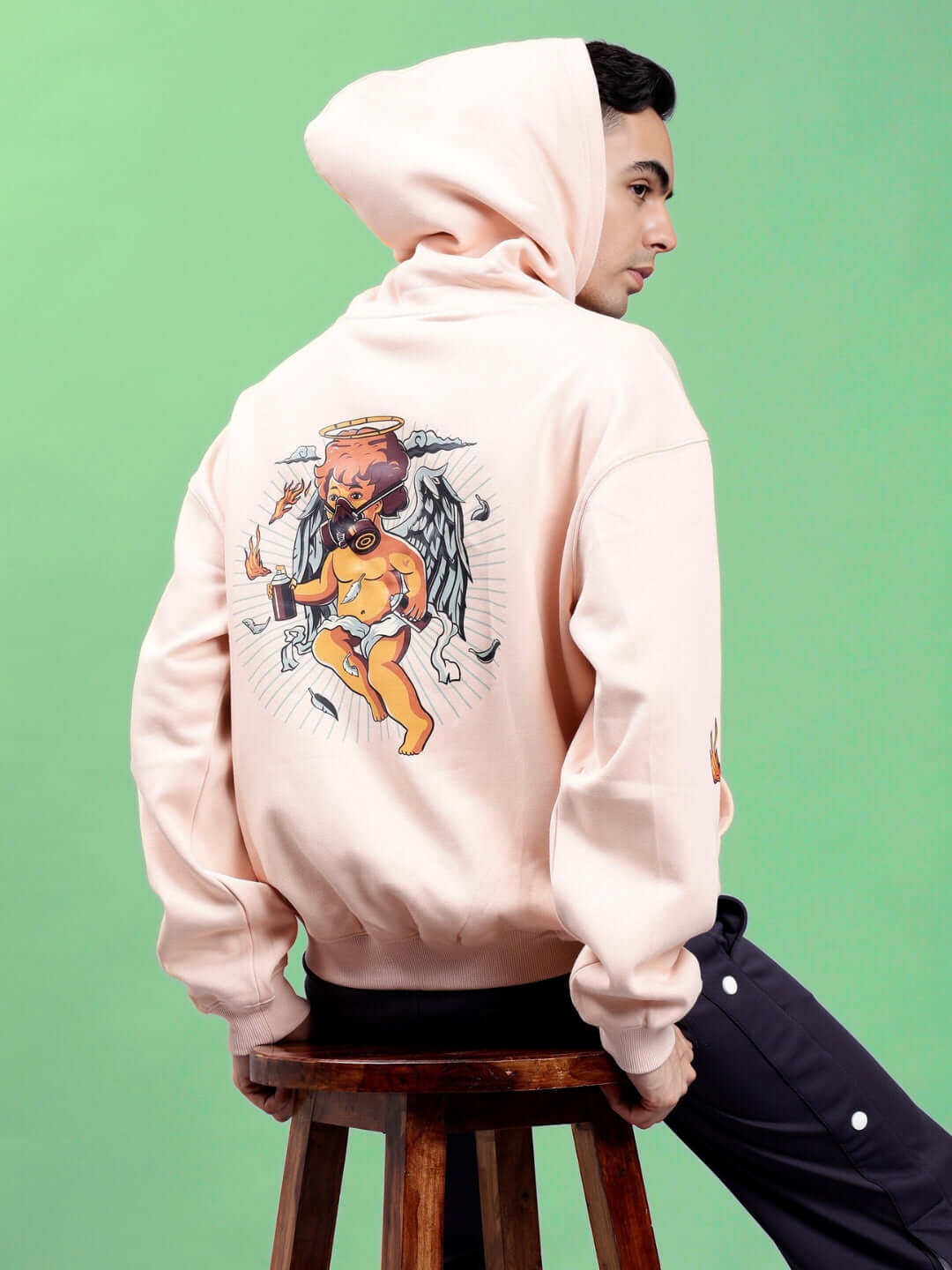 Man wearing DRIP CUPID OVERSIZED HOODIES BEIGE sitting on a stool, featuring an artistic cupid graphic on the back.