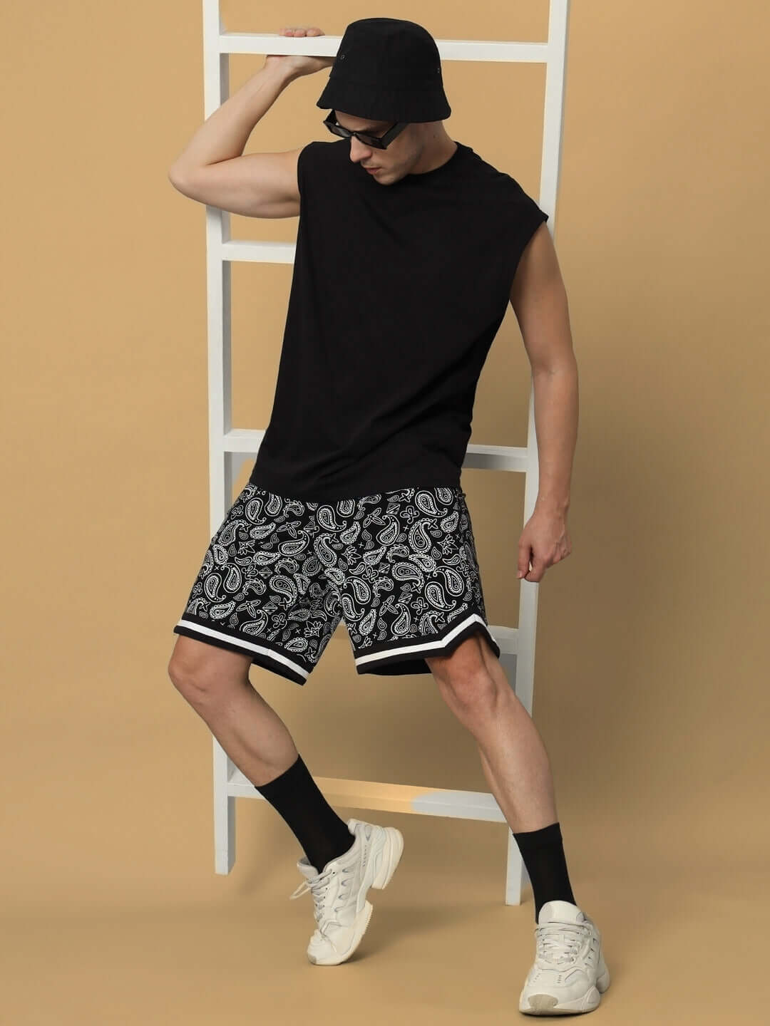 Man wearing DRIZZLE regular fit black shorts with custom size option, paired with black top, hat, and sneakers, against a beige backdrop.