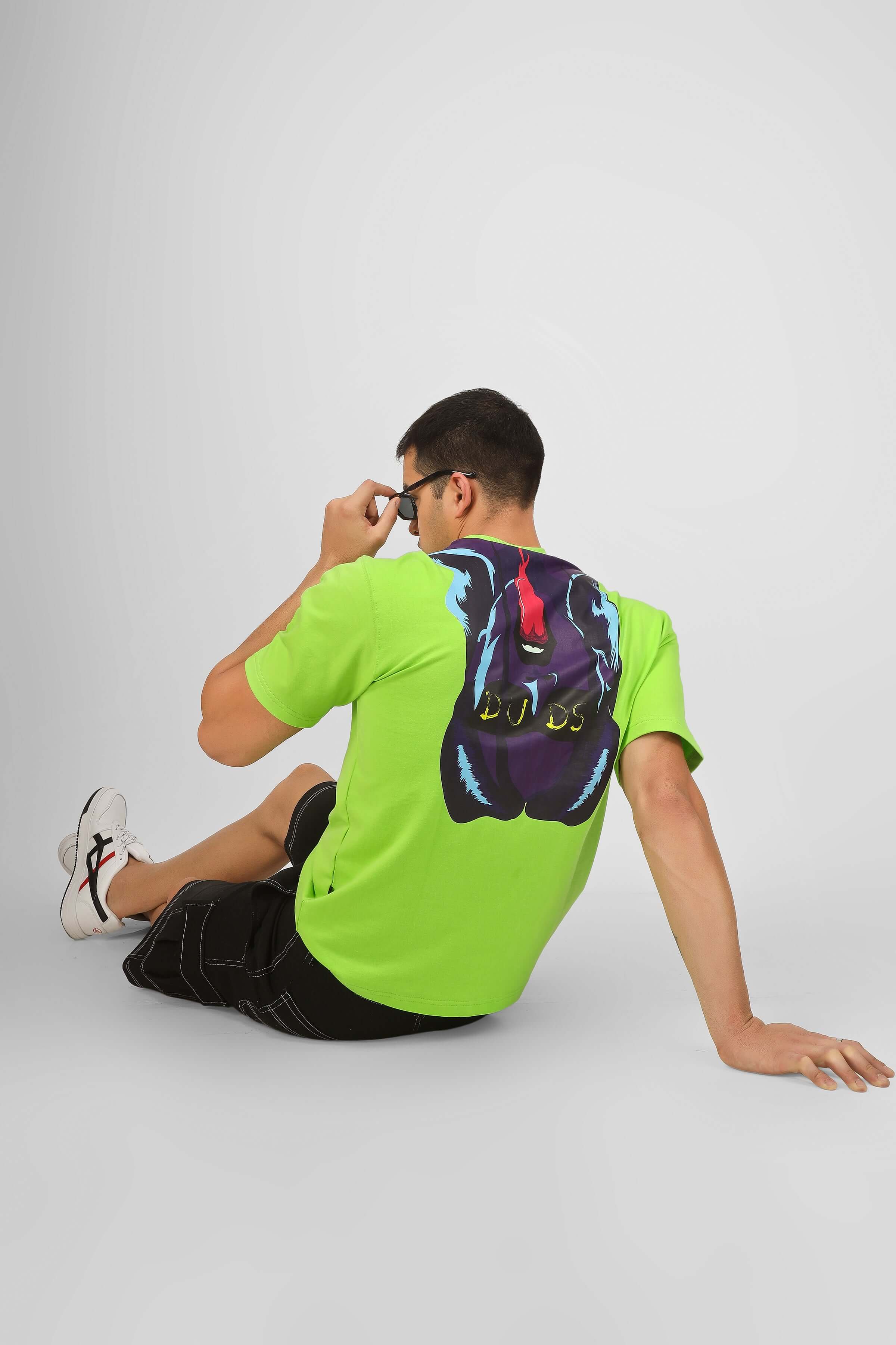 Man wearing DUDS DELIGHT oversized neon green t-shirt with graphic, sitting casually in shorts and sunglasses, showcasing back design.