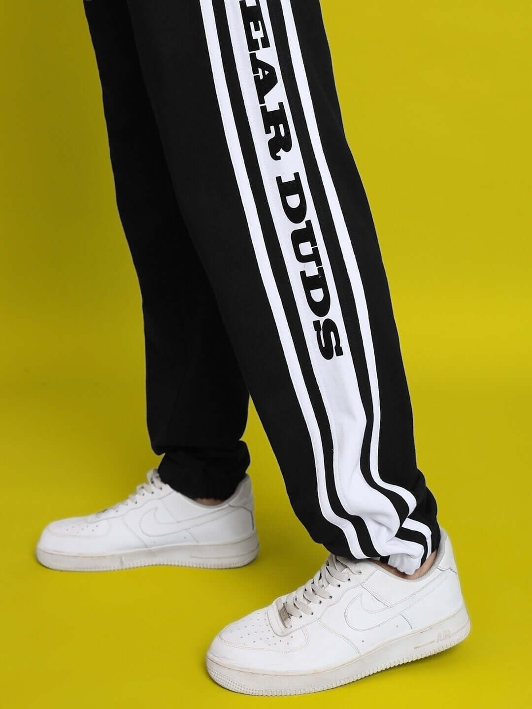 Black and white contrast panel joggers by DUDS with bold logo, worn with white sneakers on bright yellow background.