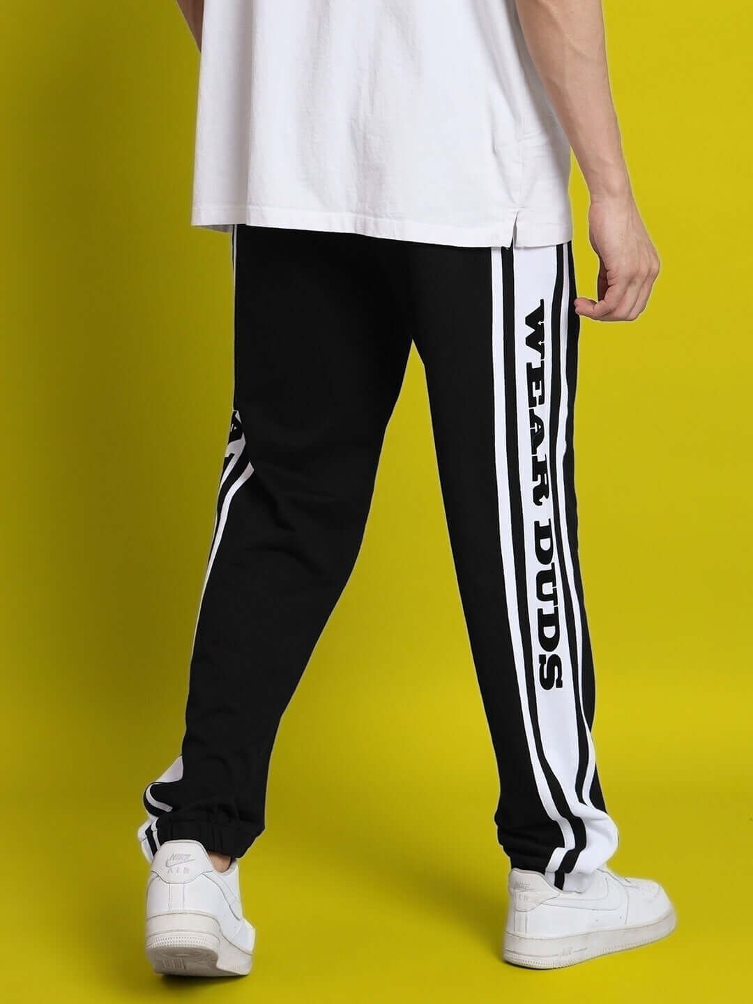 Person wearing DUDS HD contrast panel joggers in black-white with 'Wear DUDS' text, paired with white sneakers, against yellow background.