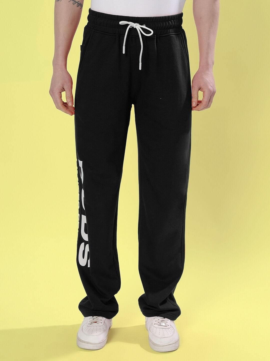 Man wearing DUDS Limited Edition black jogger pants with DUDS logo, drawstring waist, and white sneakers against a yellow background.