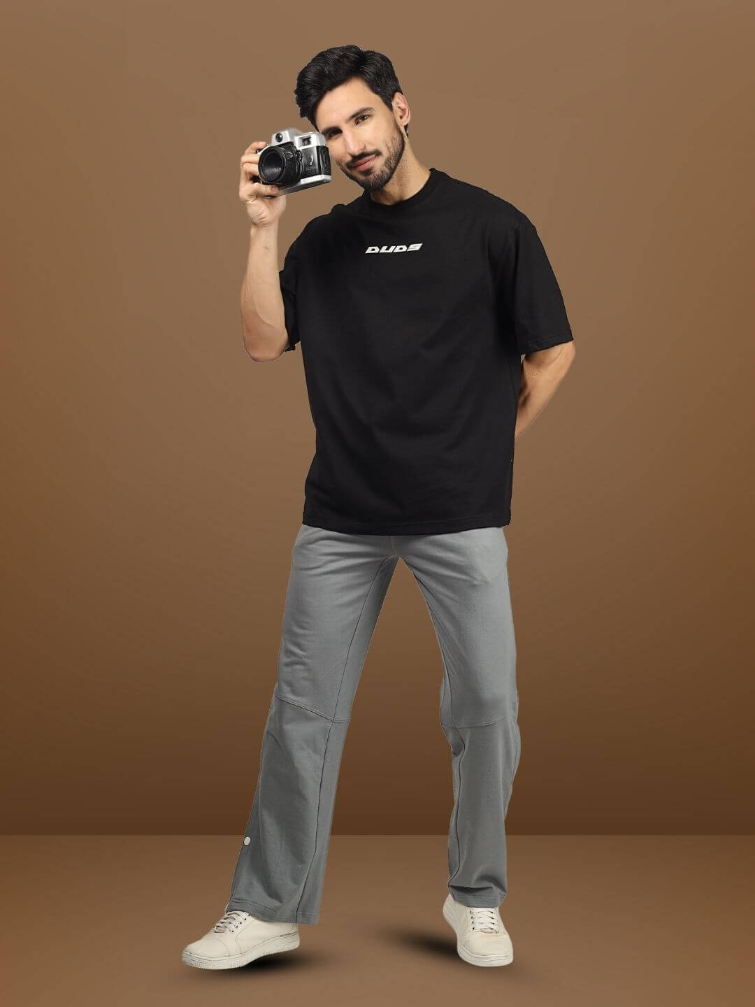 Model wearing DUDS Racer Joggers in grey paired with a black DUDS t-shirt, holding a camera.