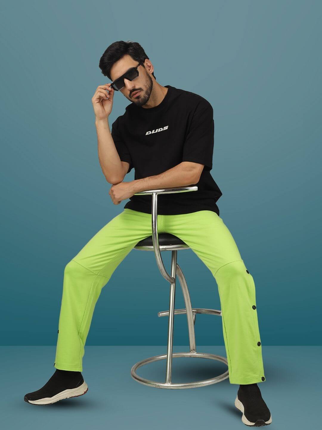 Model wearing neon green DUDS Racer Joggers seated on a stool, paired with a black t-shirt and sunglasses against a blue background.