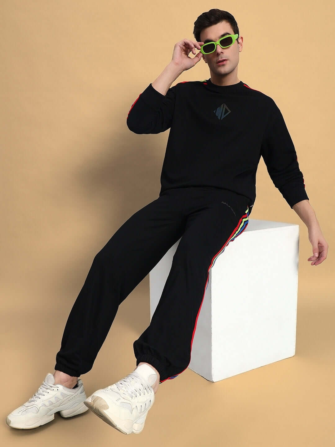 Model wearing DUDS black joggers with rainbow stripe, seated on a white block, accessorized with green sunglasses and white sneakers.