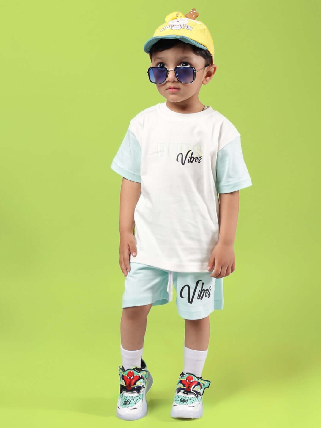 Young child modeling DUDS VIBES SHORTS FOR BOYS & GIRLS SKY BLUE with matching t-shirt and sneakers against green background.