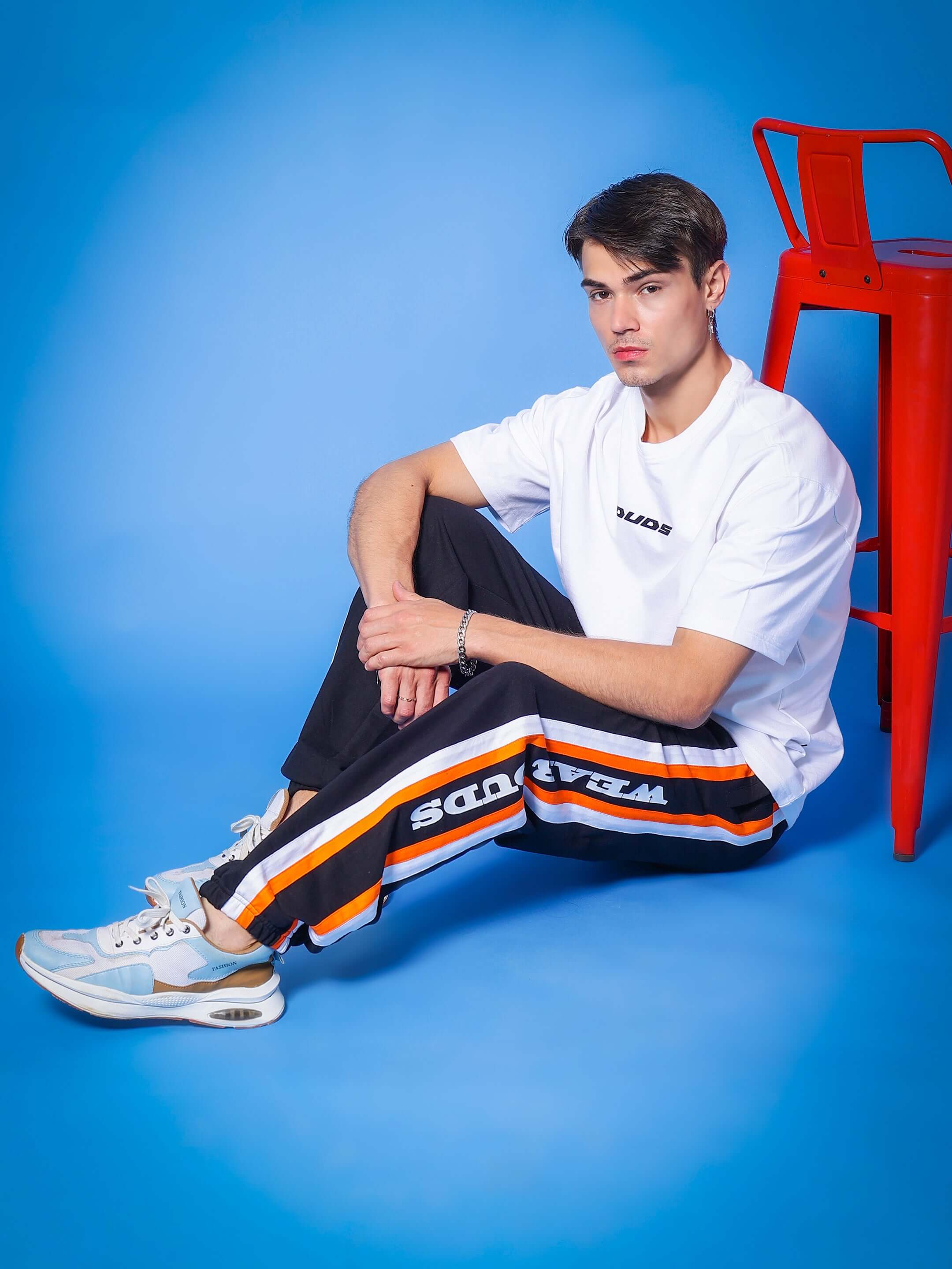Model wearing DUDS HD Bolt Joggers in black, sitting beside a red chair on a blue background.