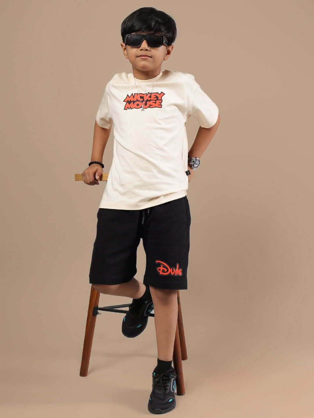 Child in DUDS beige-black Mickey Mouse co-ord on stool, wearing sunglasses, promoting stylish boys' and girls' outfit.
