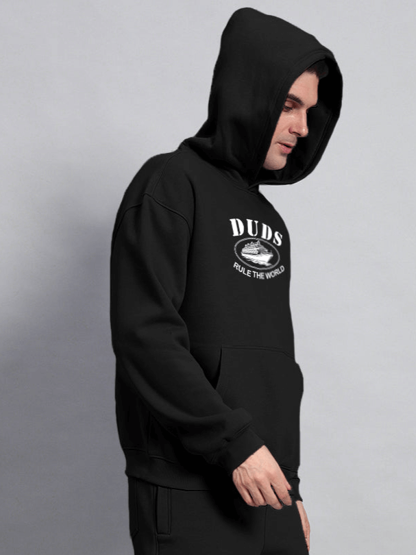 Man wearing RULE THE WORLD OVERSIZED HOODIE BLACK, showcasing streetwear style with a relaxed fit.