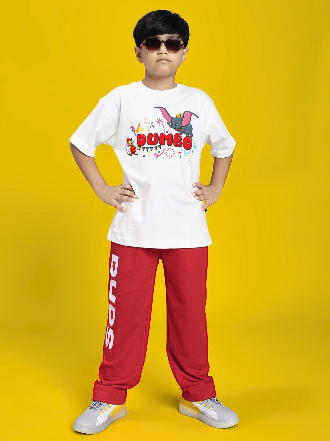 Child wearing a DUMBO T-SHIRT FOR BOYS AND GIRLS WHITE, paired with red pants, standing confidently on yellow background.