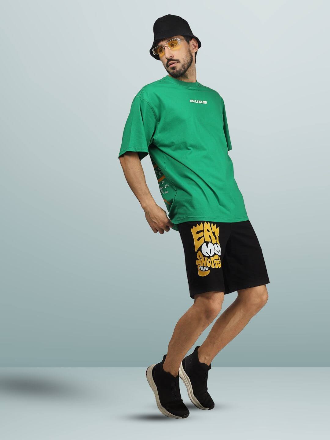 Model wearing EAT MY SHORTS Regular Fit black shorts with yellow graphic, paired with a green t-shirt, black bucket hat, and sneakers.