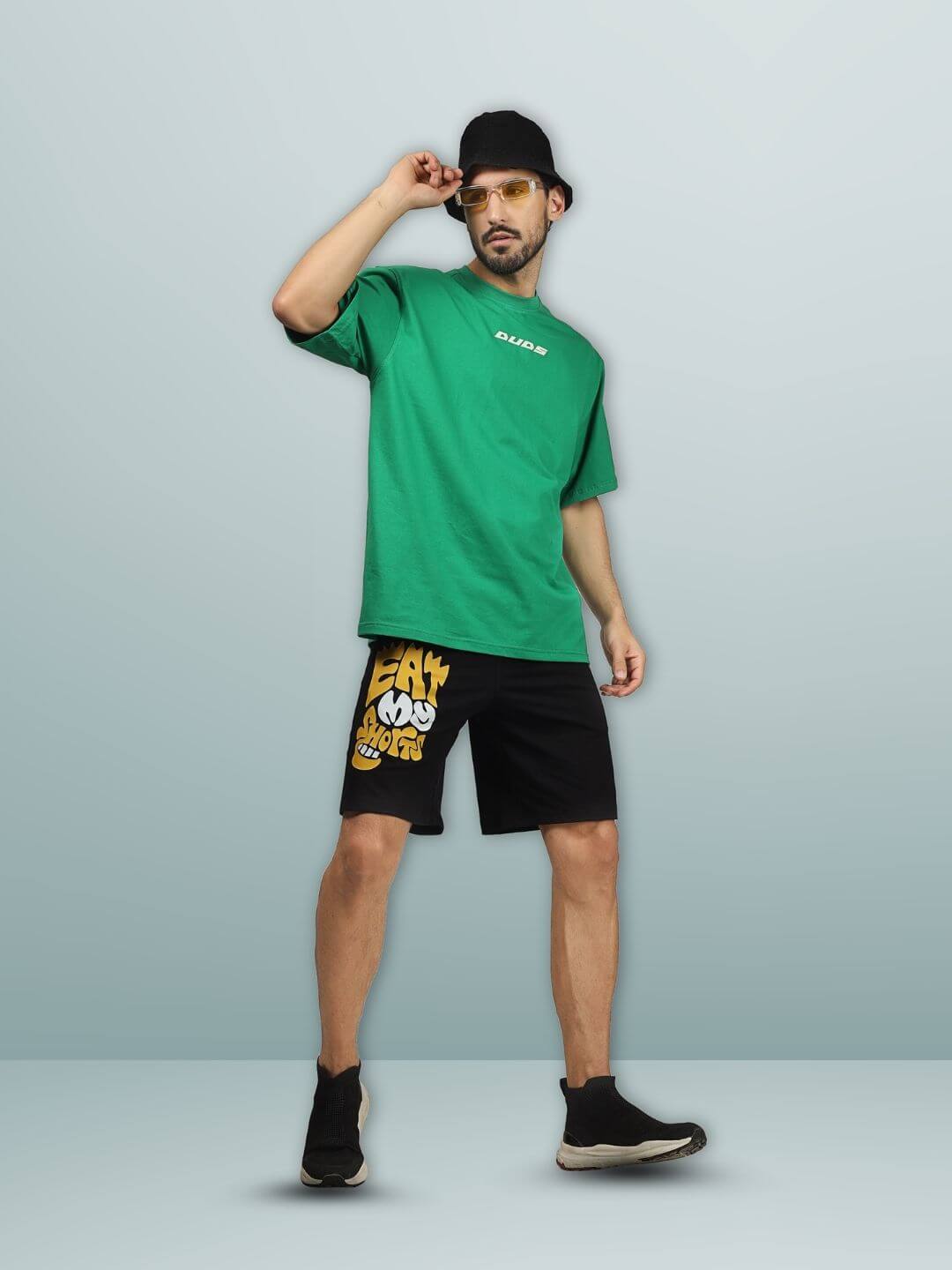 Model wearing EAT MY SHORTS regular fit black shorts with green T-shirt and black cap, showcasing casual style.