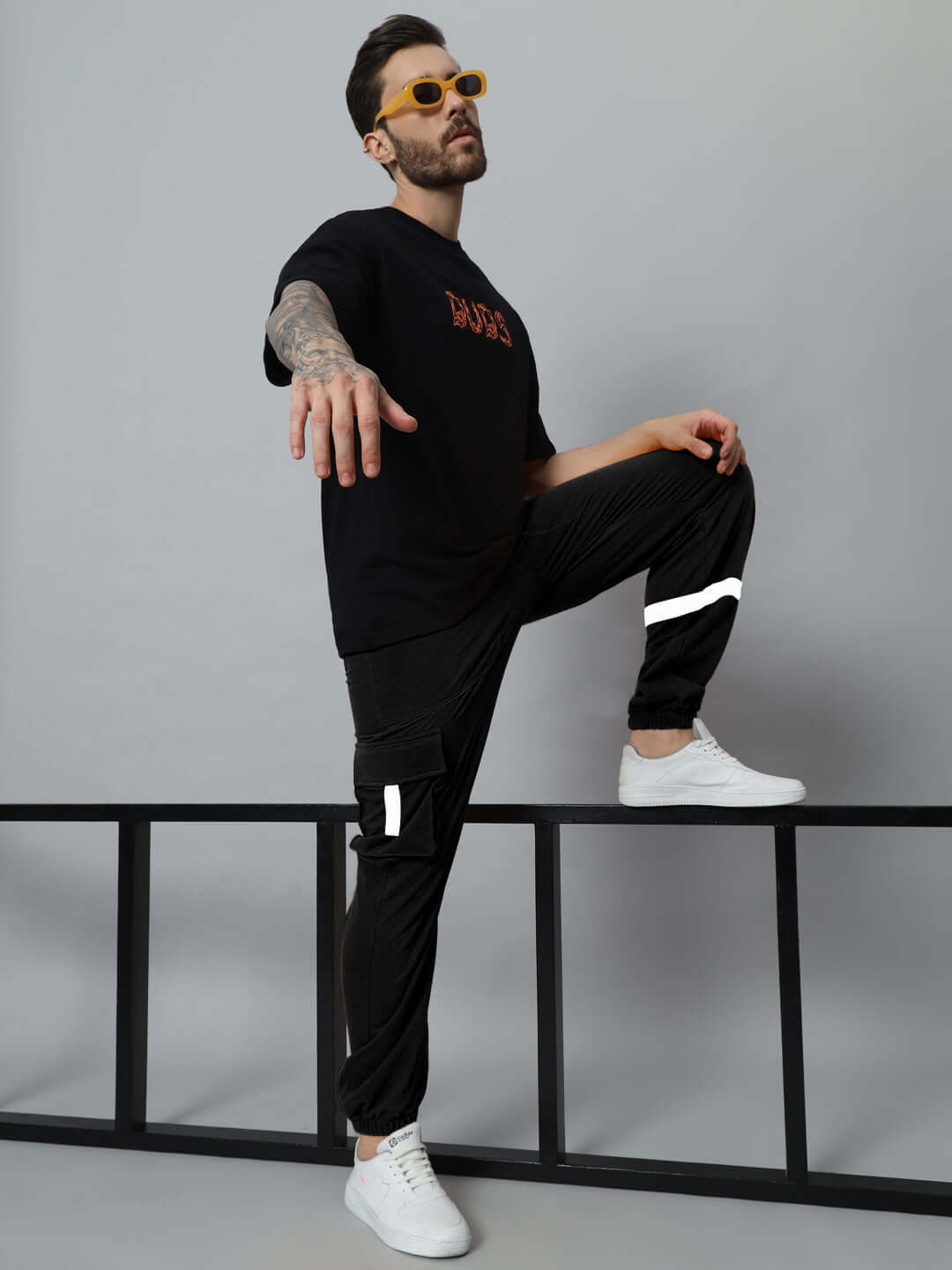 Man wearing EDIFICE relaxed fit black cargo pants and black shirt, standing confidently with one leg on bench, showcasing street style fashion.