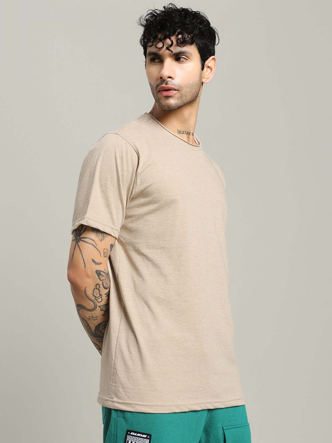 Man wearing ELEX REGULAR FIT T-SHIRT BEIGE with tattoos on arm, standing sideways in stylish pose.
