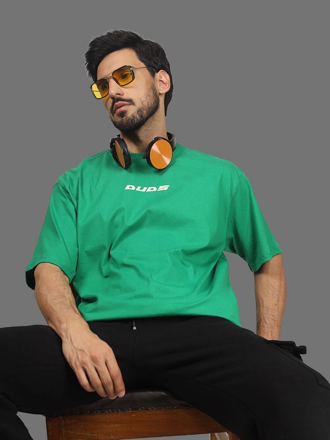Man wearing green ETCETERA oversized T-shirt by DUDS, styled with headphones and sunglasses, promoting casual fashion.