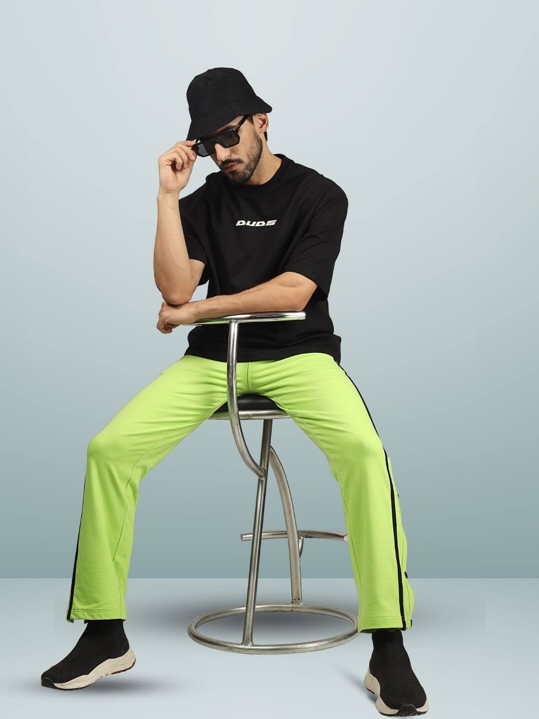 Man wearing black oversized T-shirt with "DUDS" logo seated on a stool, paired with bright green pants and hat