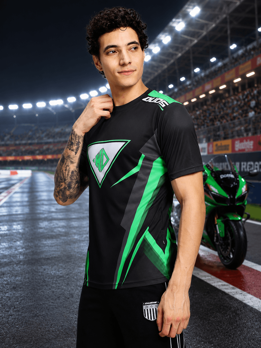APEX RACING T-SHIRT BLACK worn by a model in a racing environment with a motorcycle in the background.