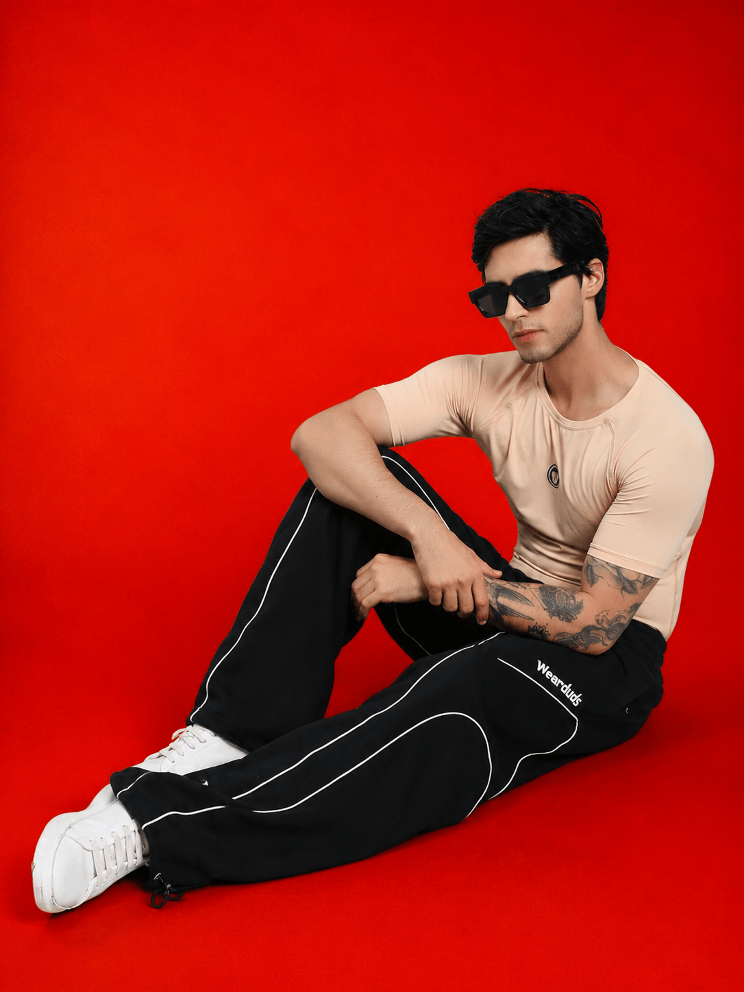 Model wearing SIGNATURE PIPING JOGGERS BLACK and a beige shirt with sunglasses on a red background.