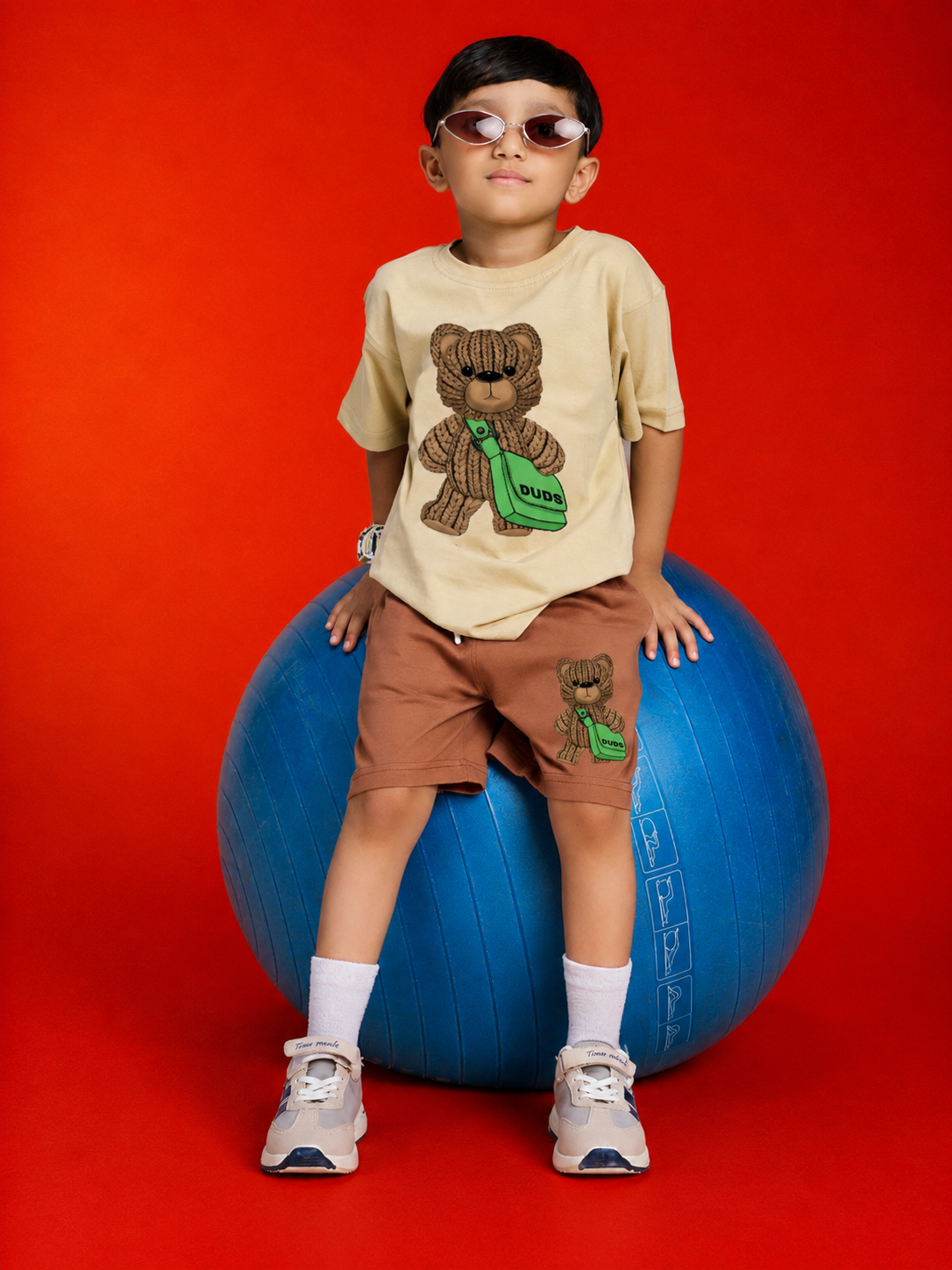 LITTLE EXPLORER TEDDY T-SHIRT FOR BOYS GIRLS BEIGE worn by a child on a blue exercise ball against a red background.