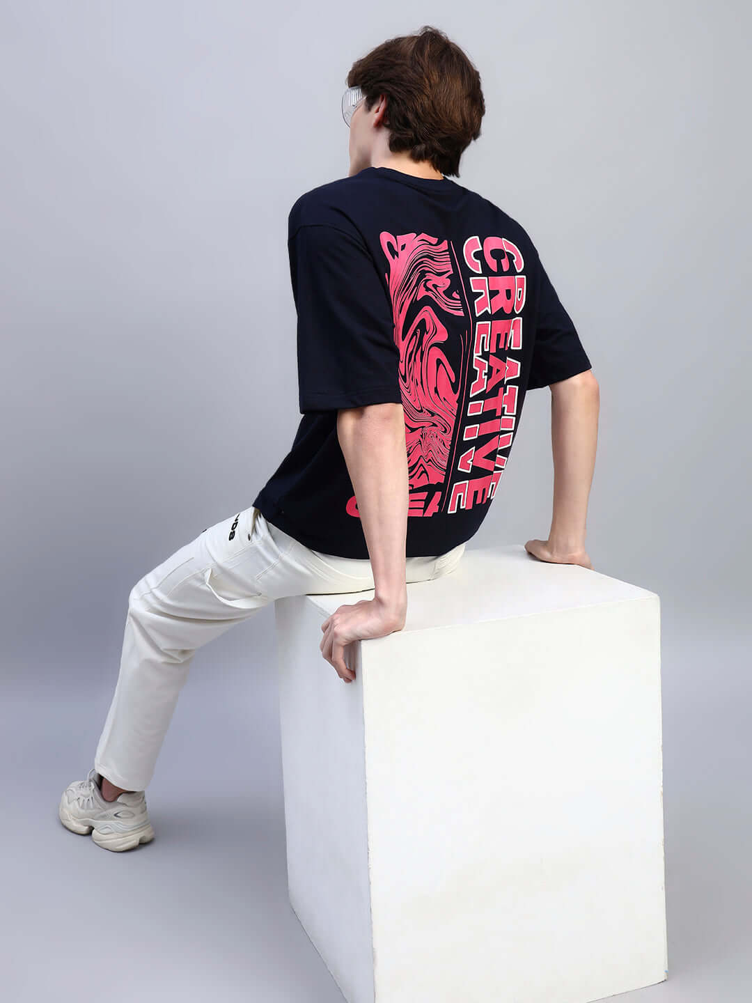 Person wearing FLORES CREATIVE PRINT OVERSIZED T-SHIRT NAVY BLUE, showing back design, sitting on white cube, casual style display.